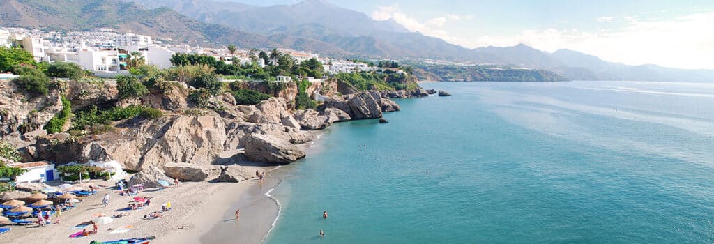 Luftaufnahme eines sonnigen Strandabschnitts mit Badegästen, umgeben von felsigen Klippen und mediterraner Architektur im Hintergrund, mit einem Blick auf das ruhige, türkisfarbene Meer und bergige Landschaften in der Ferne.